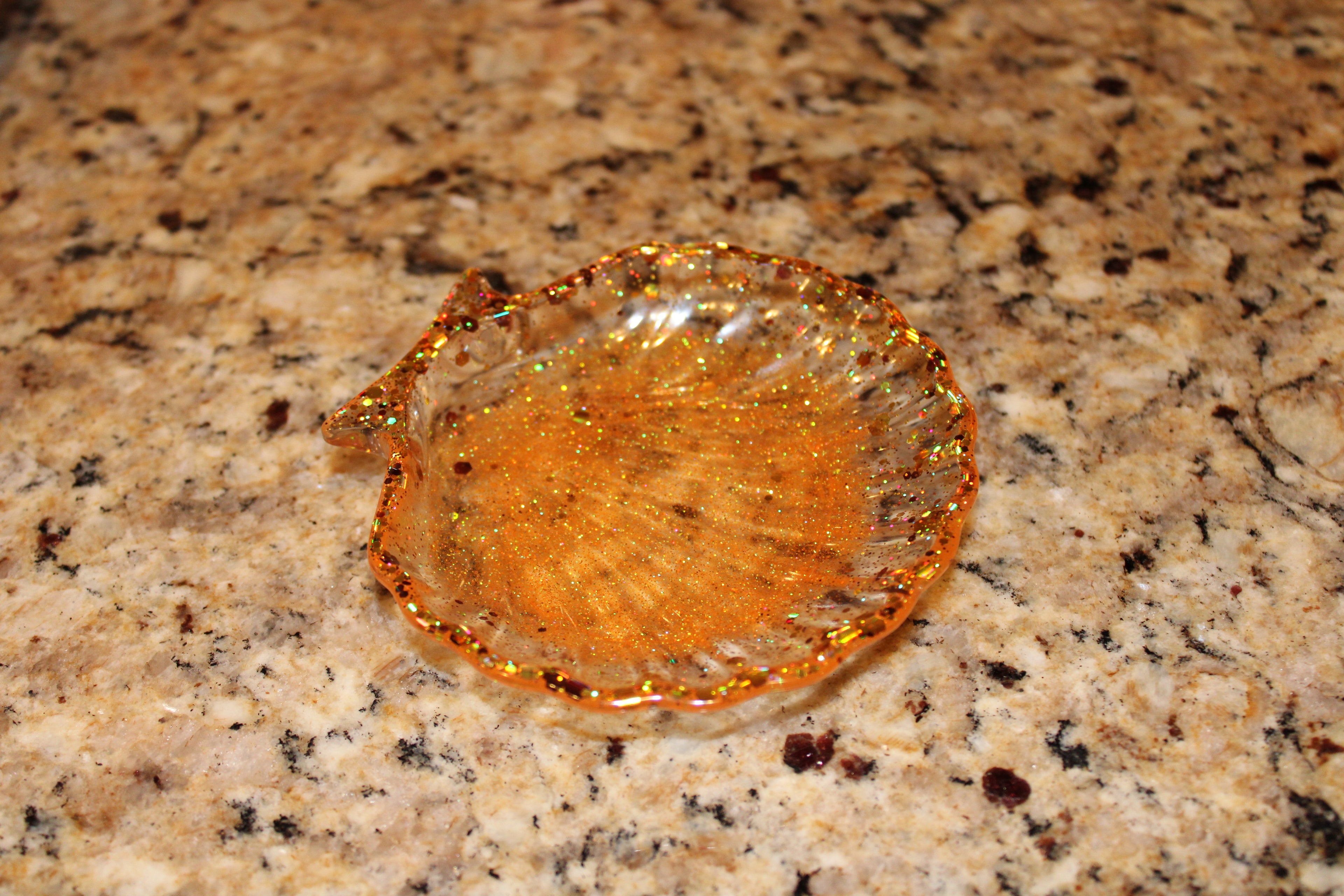 A shimmering, handmade resin seashell trinket dish sparkles with orange and gold glitter on a granite countertop.