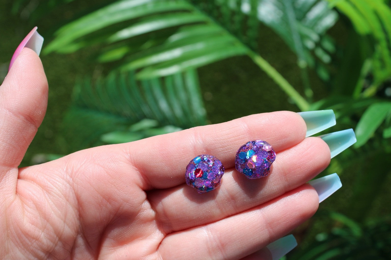 A close-up of a hand holding two small shell stud earrings with a glittery purple and iridescent blue finish, set against a blurred background of green tropical leaves.