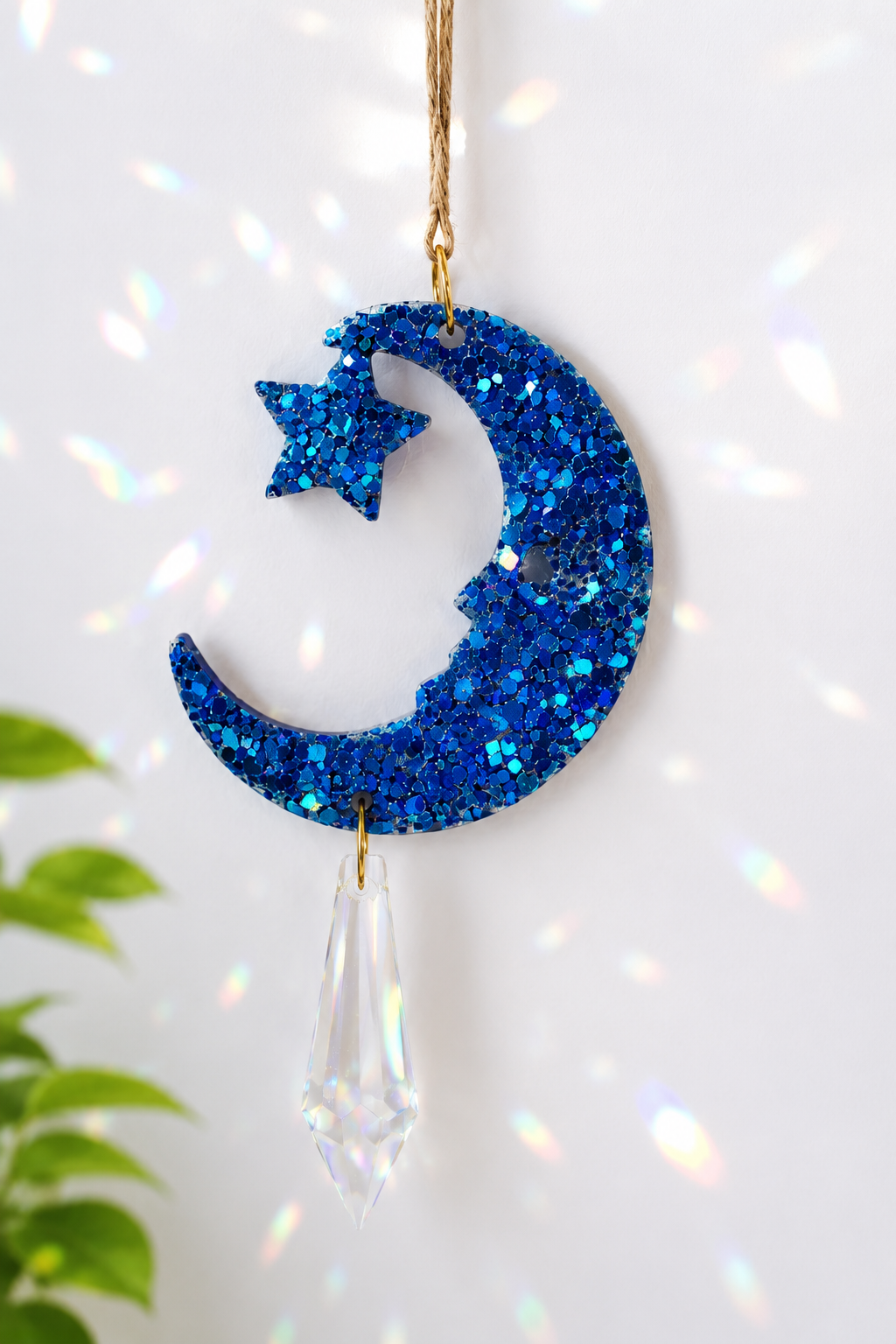 A blue glitter crescent moon and star hanging ornament with a clear faceted crystal drop, suspended by a string against a white background with iridescent light spots.