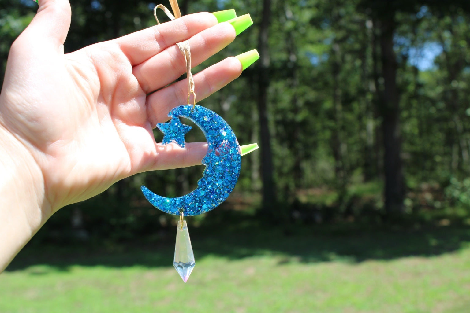 A hand with neon green fingernails holds a blue glitter crescent moon and star hanging ornament with a dangling clear crystal, set against a blurred outdoor background of trees and grass.