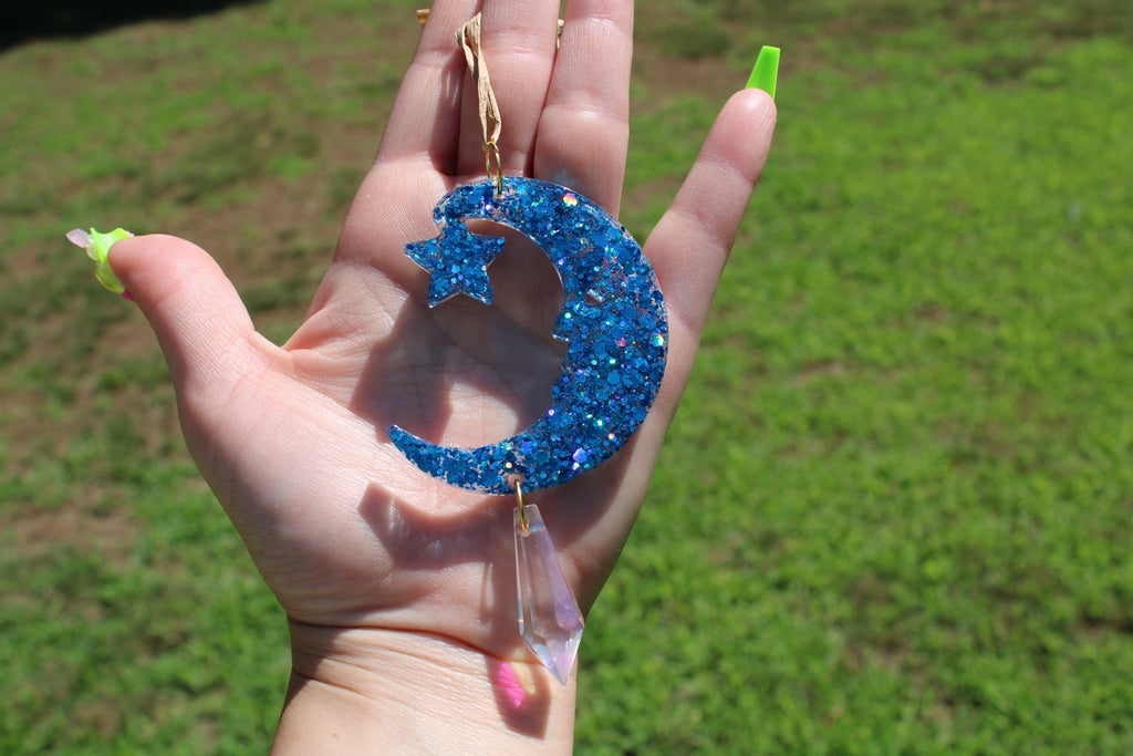 A hand holding a blue glitter crescent moon and star sun catcher outdoors with a blurred green grass background. 