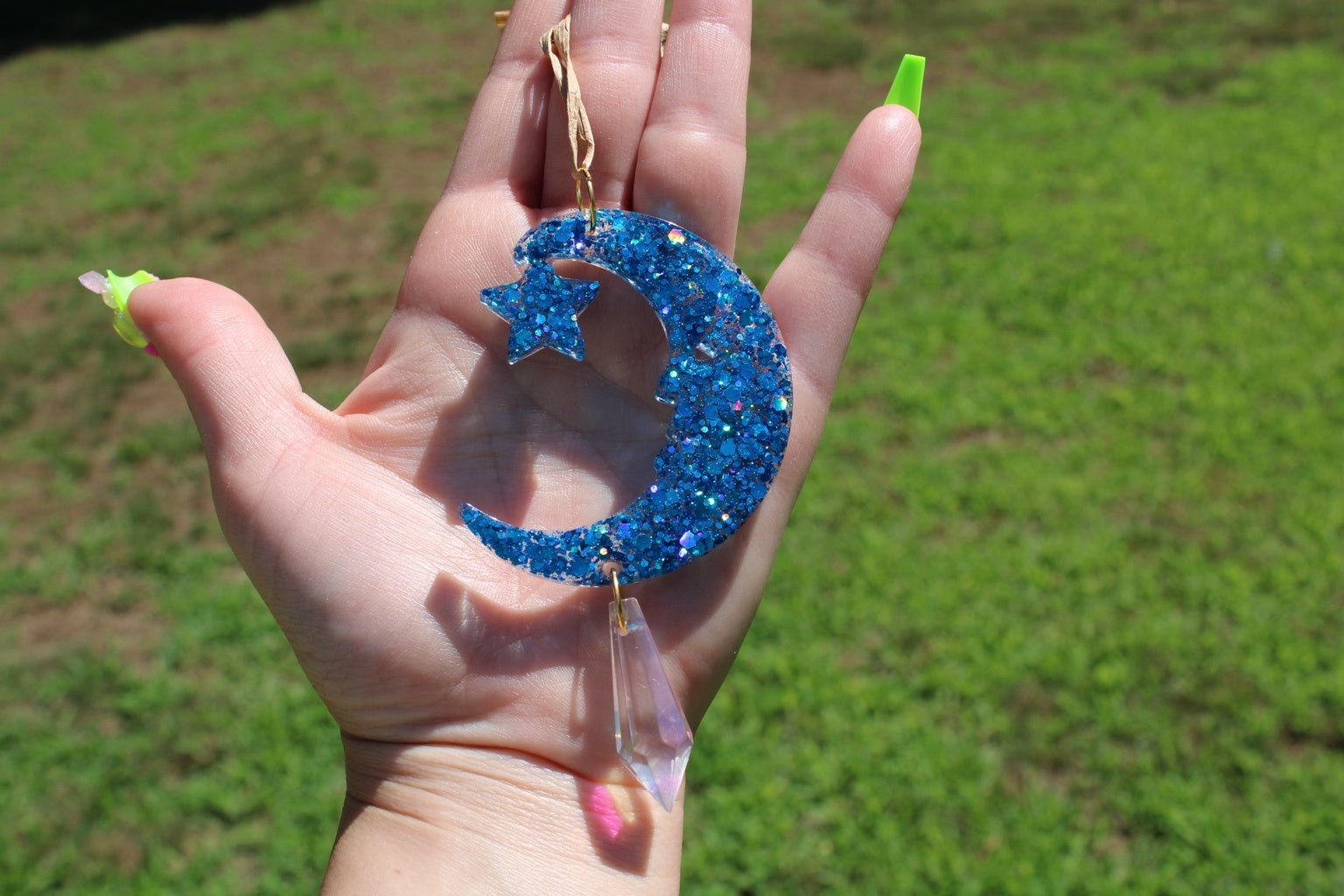 A hand holding a blue glitter crescent moon and star sun catcher outdoors with a blurred green grass background. 