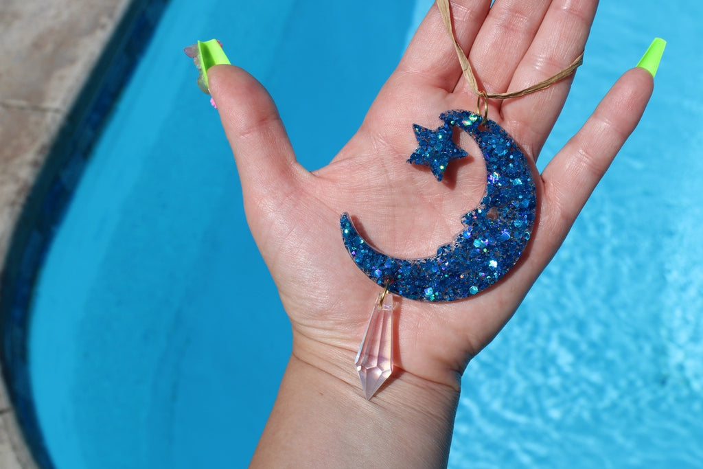 A glittery blue crescent moon and star pendant sun catcher held in a hand, with a dangling crystal. The item is shown against a bright blue swimming pool background.