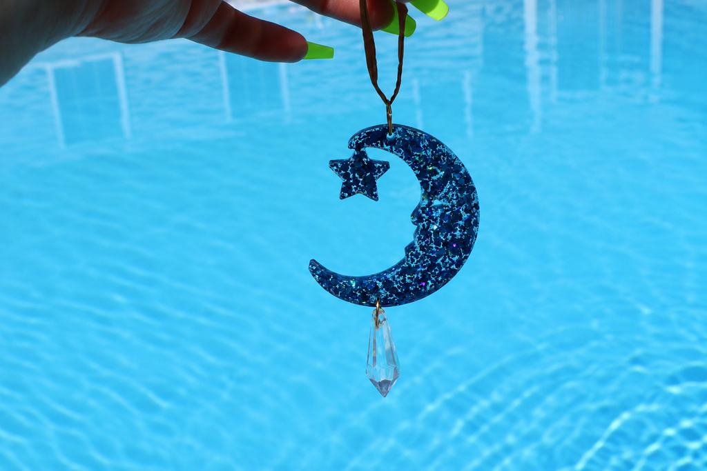 A hand with neon green nails holds a dark blue glitter crescent moon and star sun catcher with a hanging clear crystal, set against a bright blue swimming pool background.