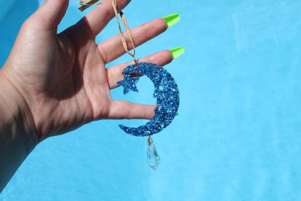 A hand with neon green fingernails holds a blue glitter crescent moon and star hanging ornament with a clear teardrop charm against a bright blue background.
