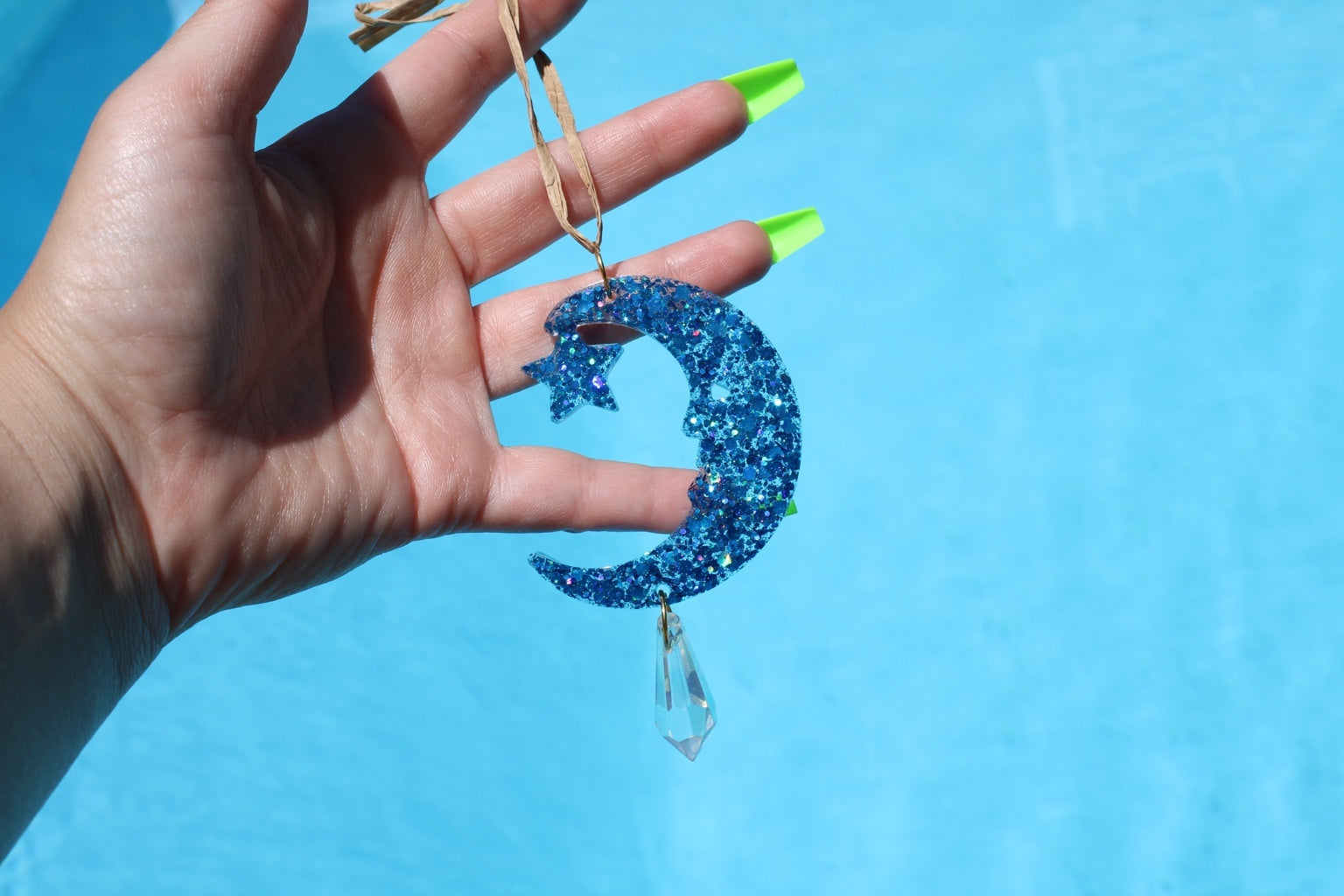 A hand with neon green fingernails holds a blue glitter crescent moon and star hanging ornament with a clear teardrop charm against a bright blue background.