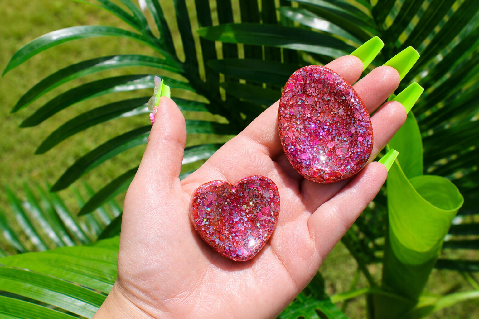 A hand with bright green fingernails holds two glittery heart-shaped fidget tokens in front of lush green palm leaves outdoors.