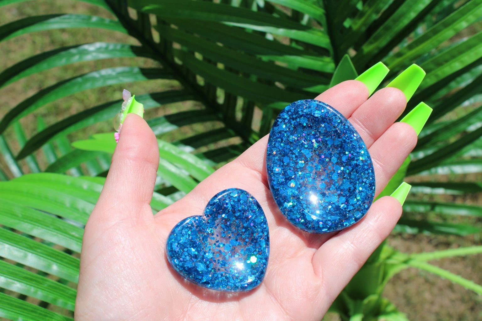 A hand with bright green manicured nails holds two glittery blue resin anxiety tokens, one heart-shaped and one oval, against a background of green palm leaves.