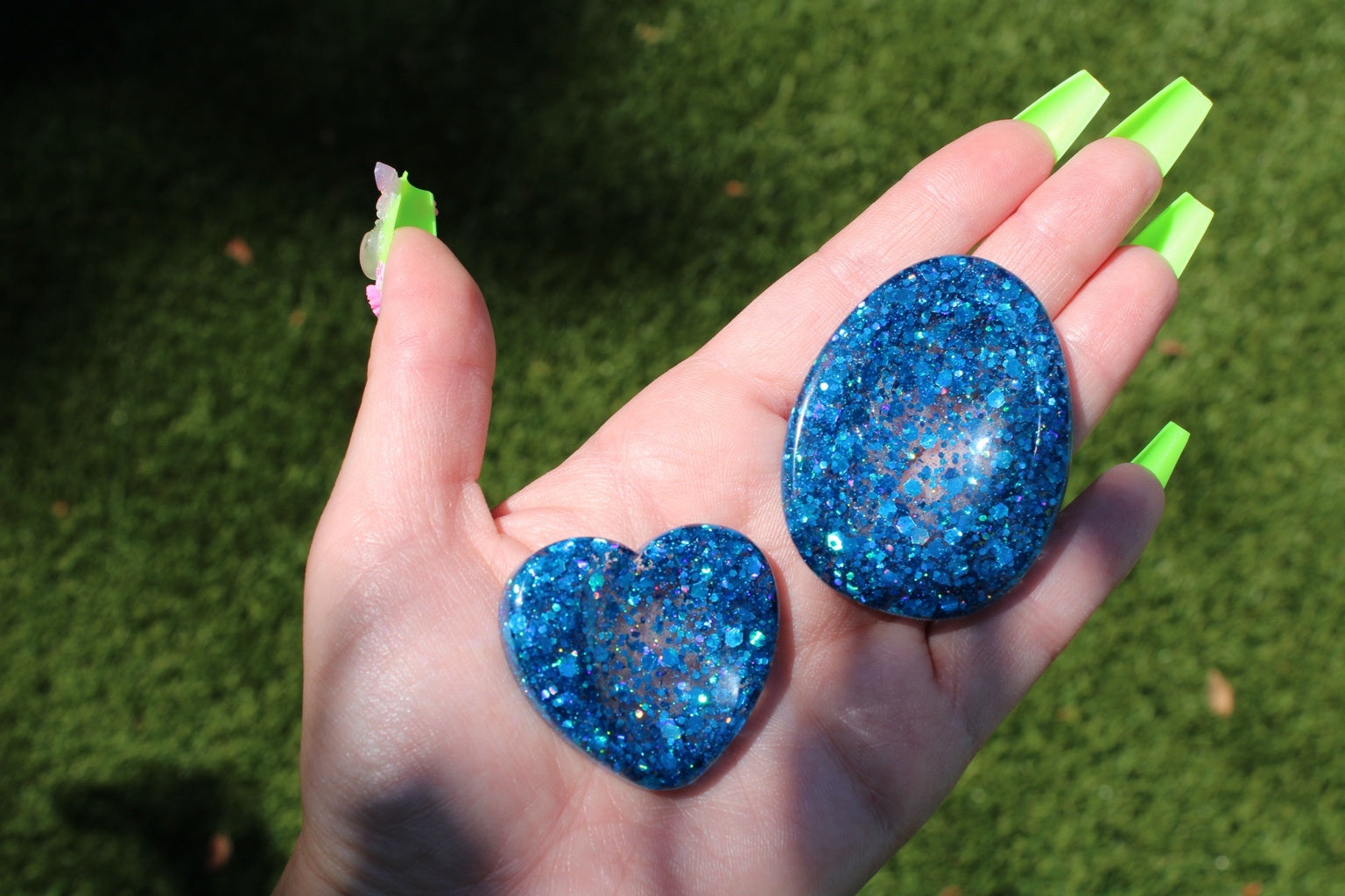 A hand with bright neon green nails holds two blue anxiety tokens against a blurred green grass background.