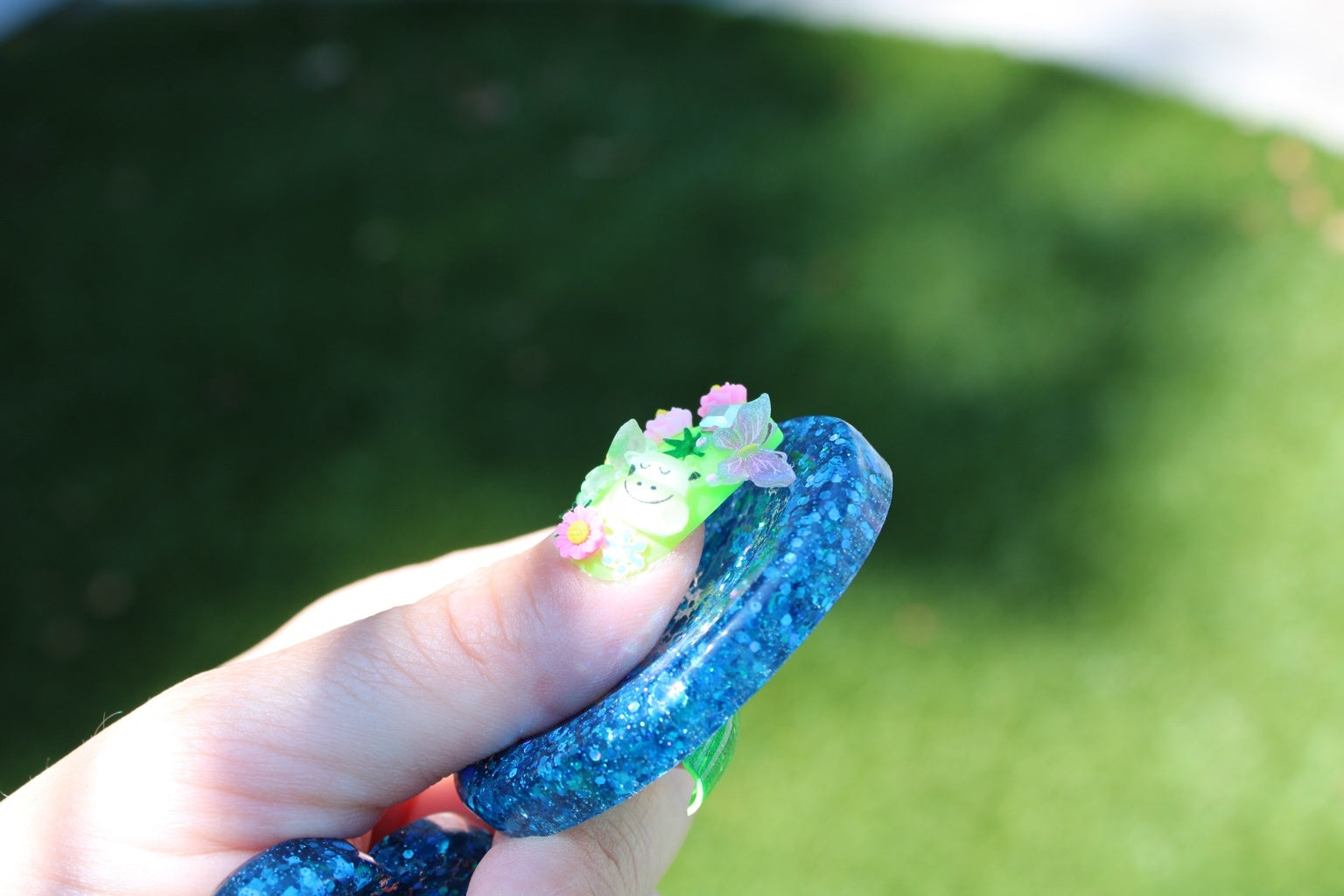 A hand holding a blue glittery oval shaped anxiety fidget token, set against a blurred green grass background.