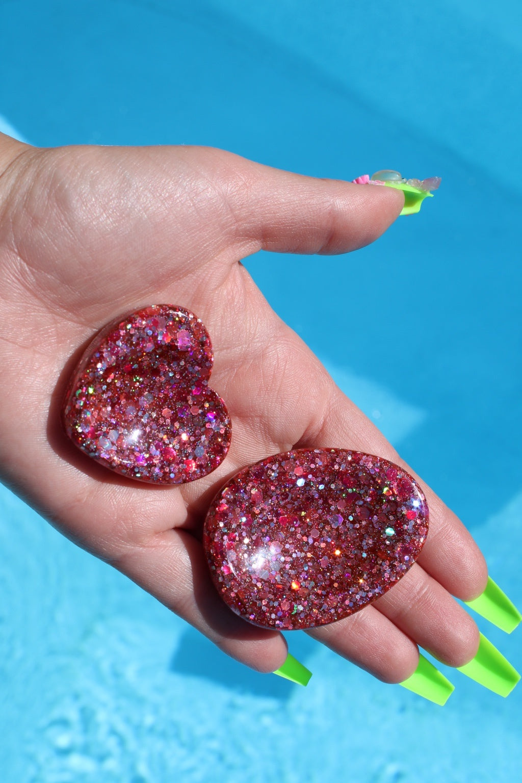A hand with neon green fingernails holds two glittery pink fidget tokens against a bright blue pool background.