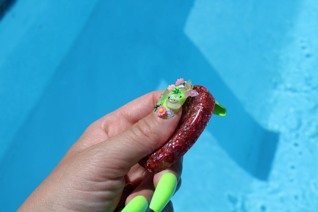 A hand with neon green nails holds a small, glittery pink anxiety token, set against a bright blue swimming pool background.