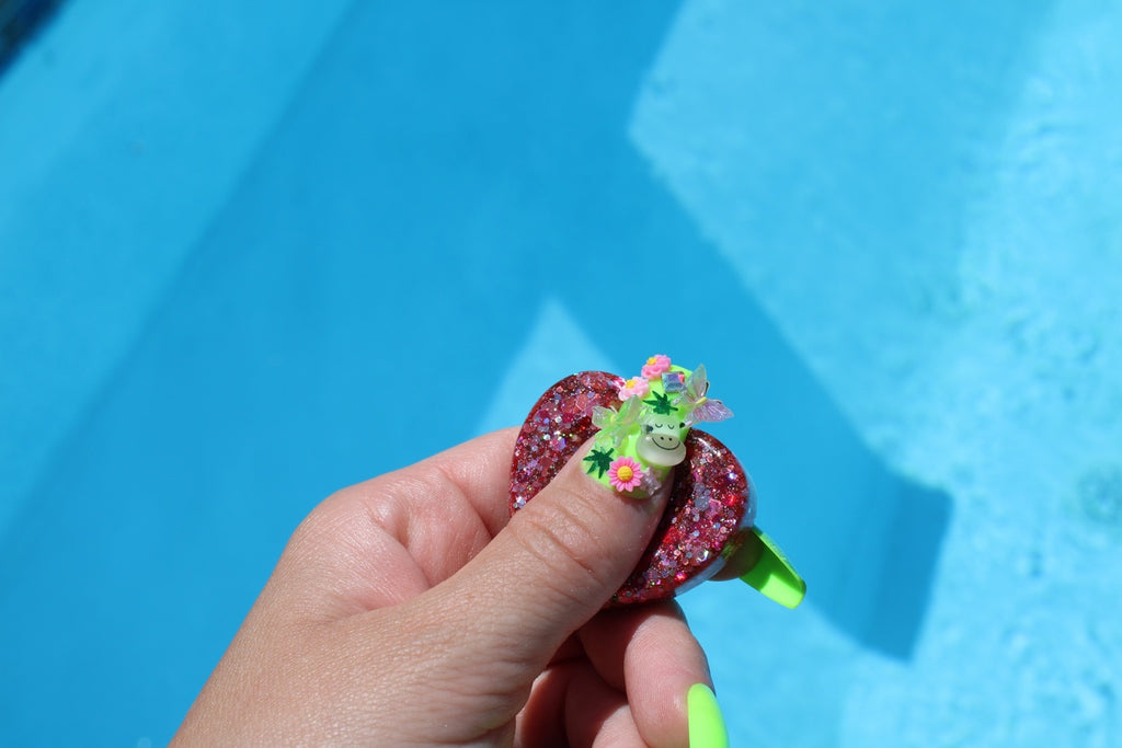 A hand holding a small, glittery pink heart-shaped fidget token, set against a bright blue swimming pool background.