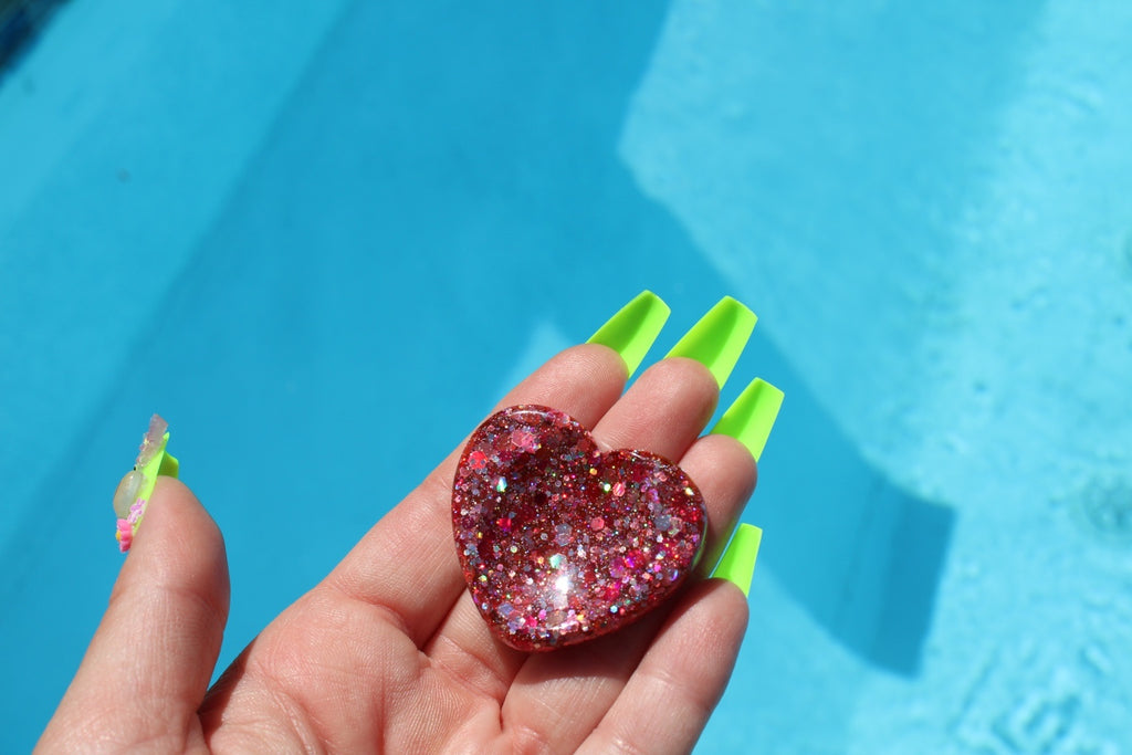 A hand with bright neon green fingernails holds a small, glittery pink heart-shaped fidget token against a bright blue swimming pool background.