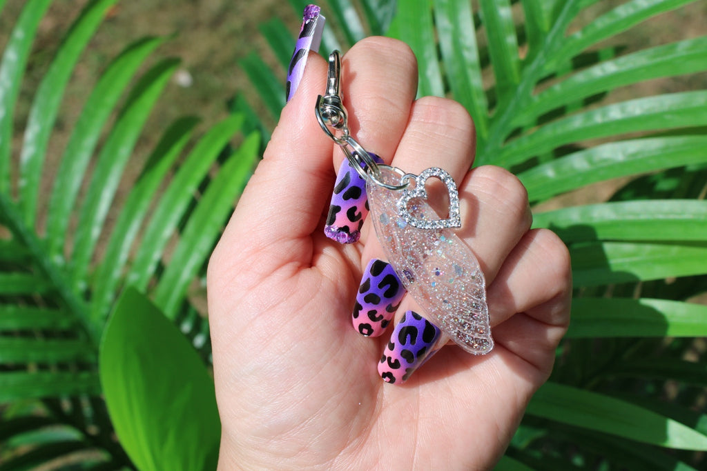 A hand with purple and pink leopard-print nails holds a clear glittery resin wing keychain with rhinestone heart, set against a background of green palm leaves.