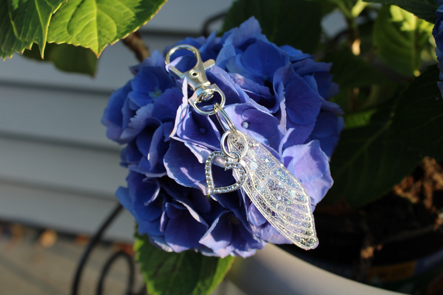 A silver-toned metal keychain with a wing-shaped charm and a small heart charm, shown hanging from a blue hydrangea flower outdoors.