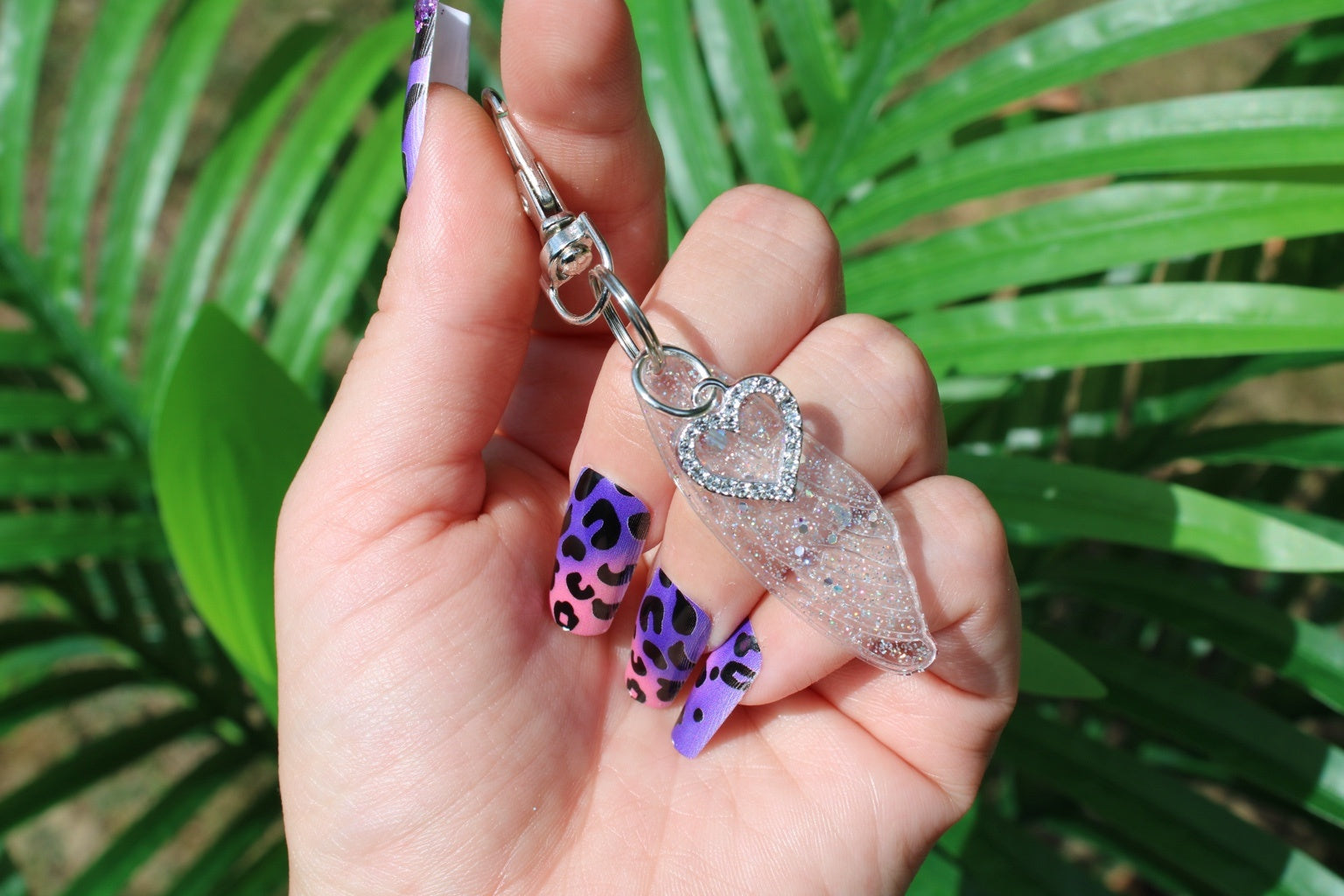 A hand with purple and black leopard-print nails holds a sequin wing keychain or bag charm with a rhinestone heart, set against a background of green palm leaves.