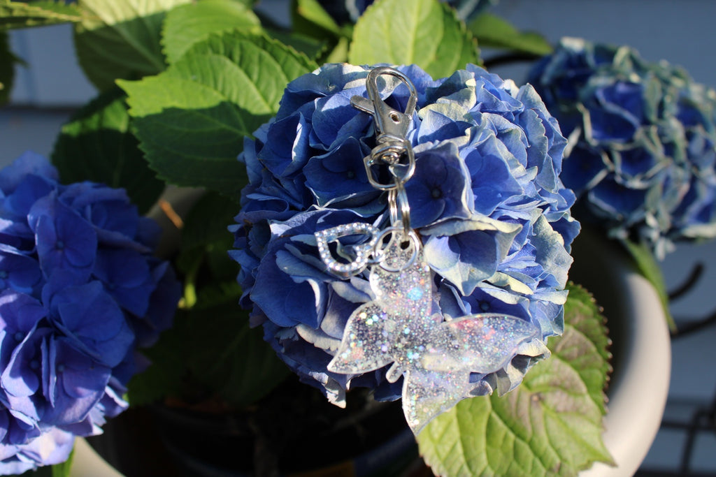 A close-up of a blue hydrangea flower with a silver-toned keychain featuring a glittery butterfly and rhinestone heart charm.