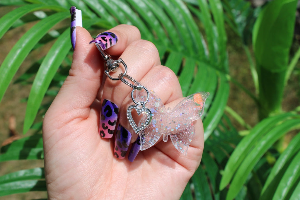 A hand with purple and black patterned nail art holds a keychain featuring a clear glitter butterfly charm and a heart-shaped charm, set against a background of green tropical leaves.