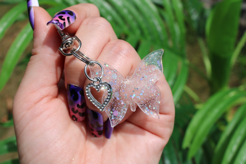 A hand with purple and black patterned nails holds a keychain featuring a clear glitter butterfly charm and a heart charm with rhinestones, set against a blurred background of green leaves.