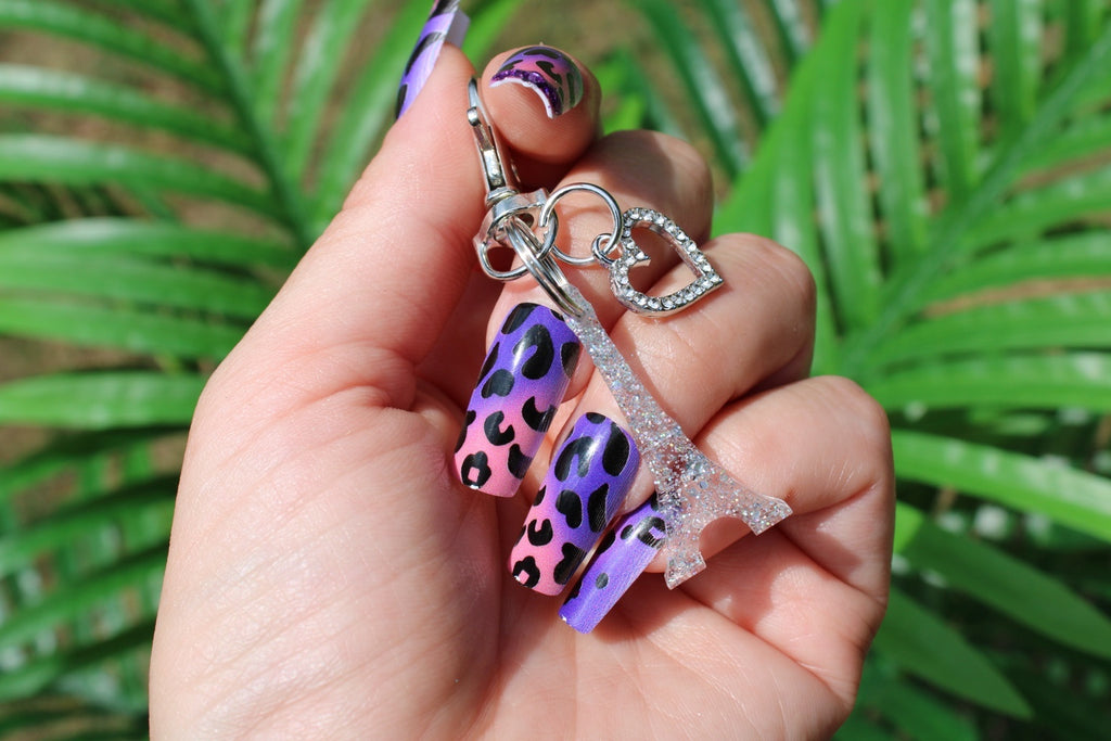 A hand with purple and pink leopard-print nail art holds a glittery Eiffel Tower charm and a rhinestone heart charm, with green palm leaves in the background.