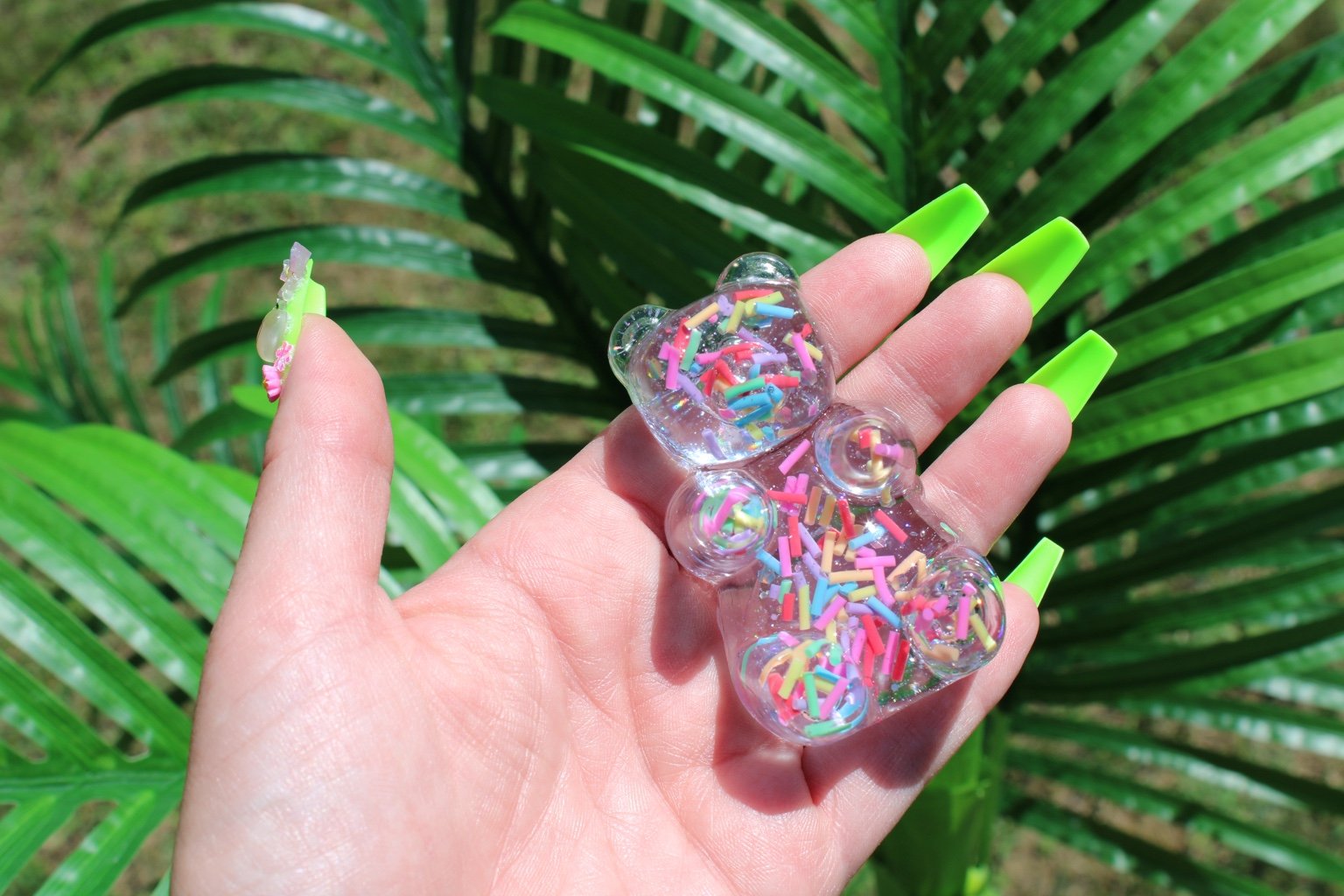 A hand with bright neon green nails holds a small clear bear-shaped resin figurine filled with colorful sprinkles, set against a background of green palm leaves.