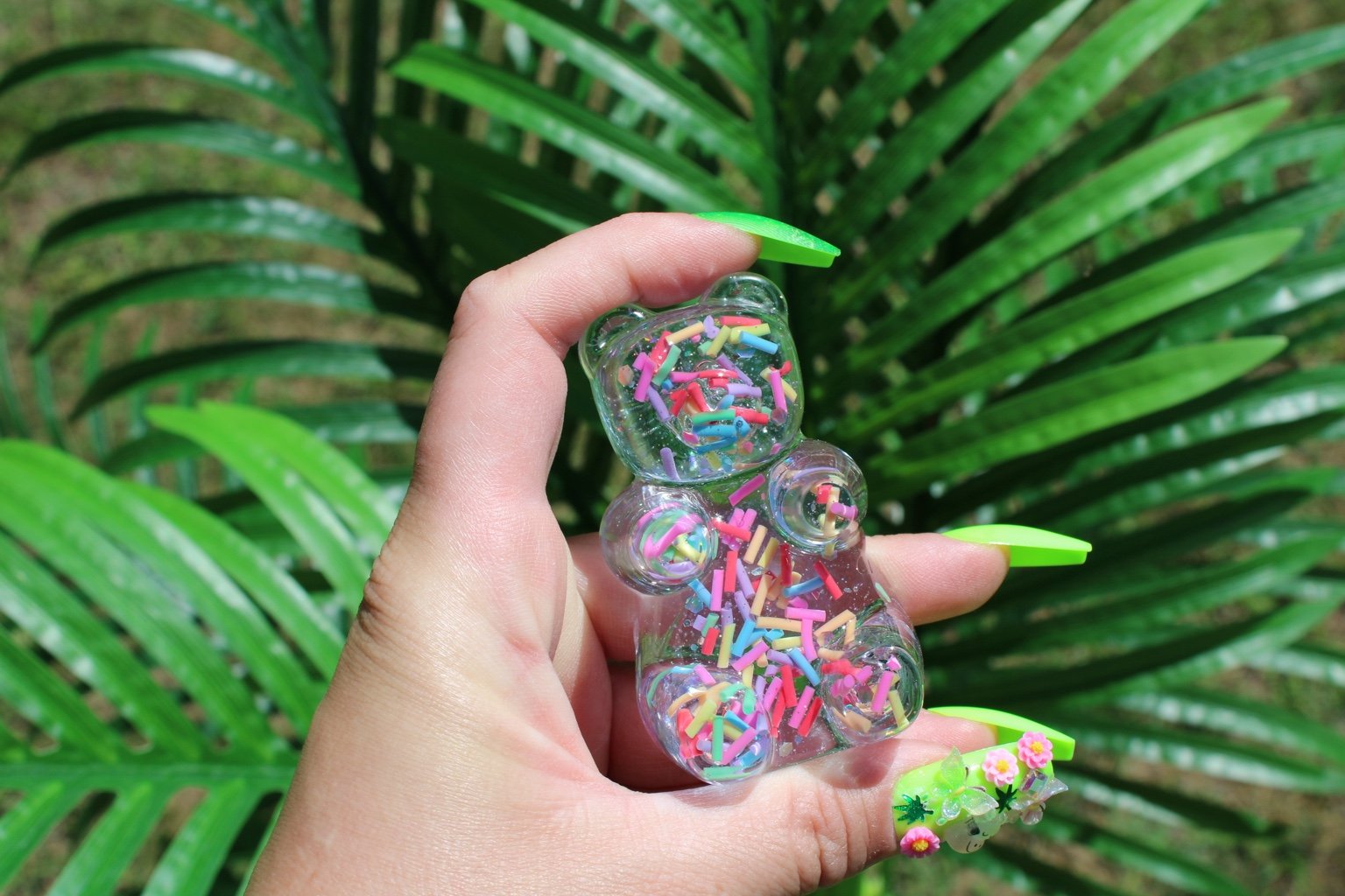 A hand with bright green nails holds a small clear bear-shaped figurine with colorful sprinkles, set against a background of large green palm leaves.