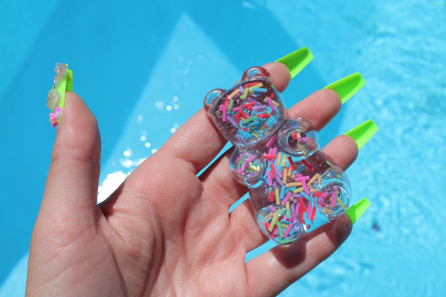 A hand with bright green nails holds a resin teddy-bear filled with multicolored sprinkles against a bright blue swimming pool background.