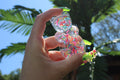 A hand holding a clear, bear-shaped resin sprinkle filled figurine, set against a sunny outdoor background of palm trees and blue sky.