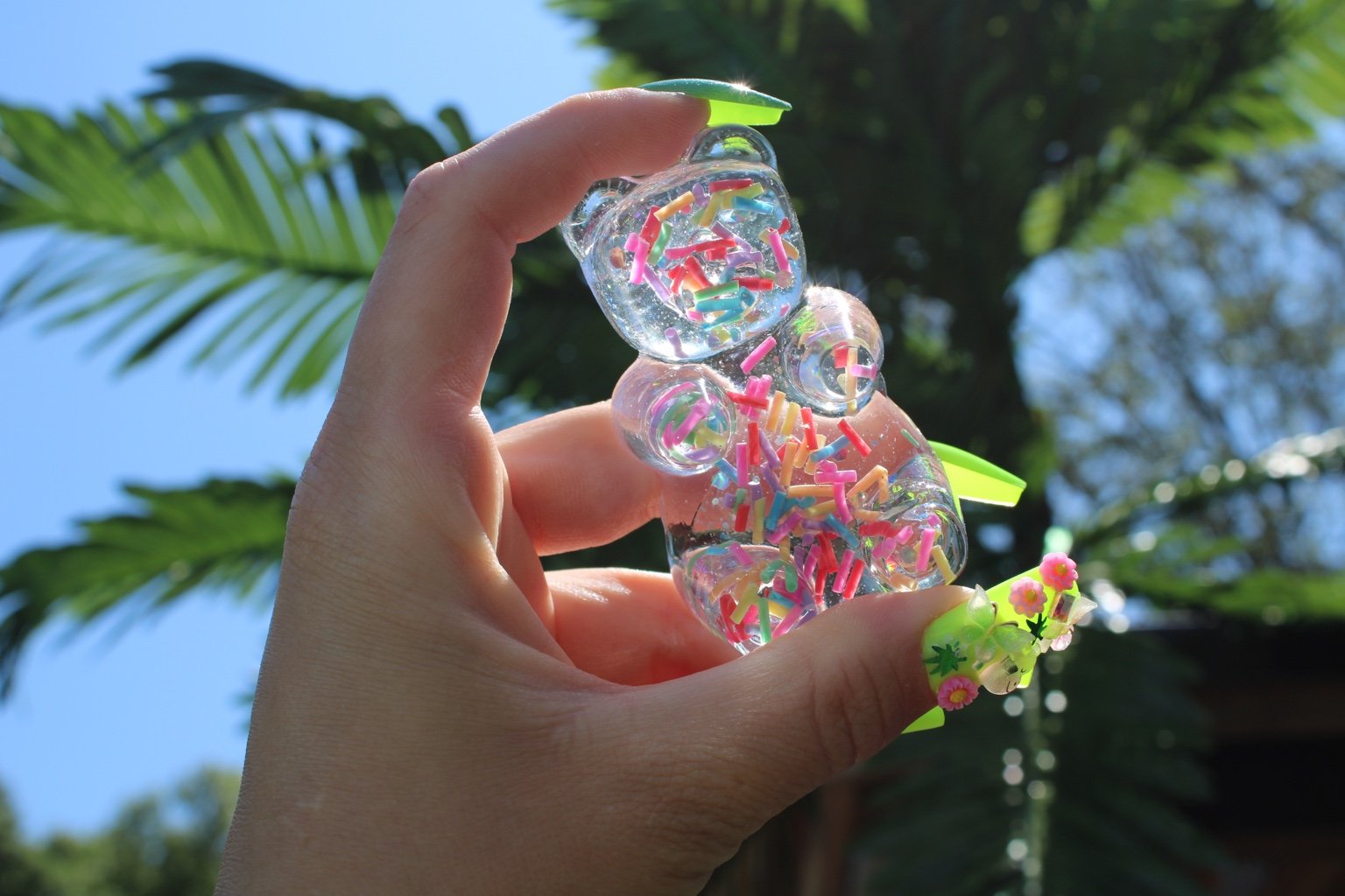 A hand holding a clear, bear-shaped resin sprinkle filled figurine, set against a sunny outdoor background of palm trees and blue sky.