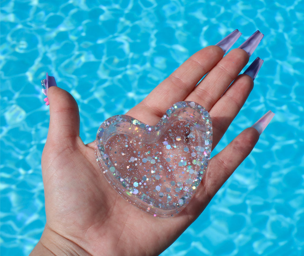 Hand holding a glittery heart-shaped object with a blue water background