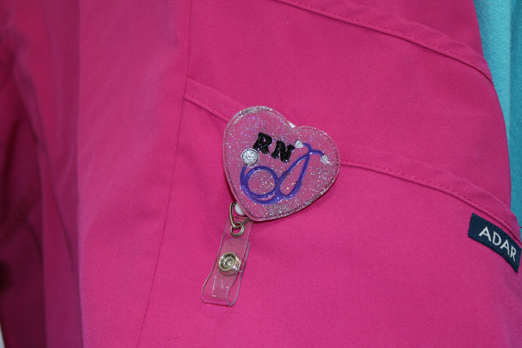 A registered nurse's pink scrub top features a glittery heart-shaped RN badge reel with a stethoscope design.