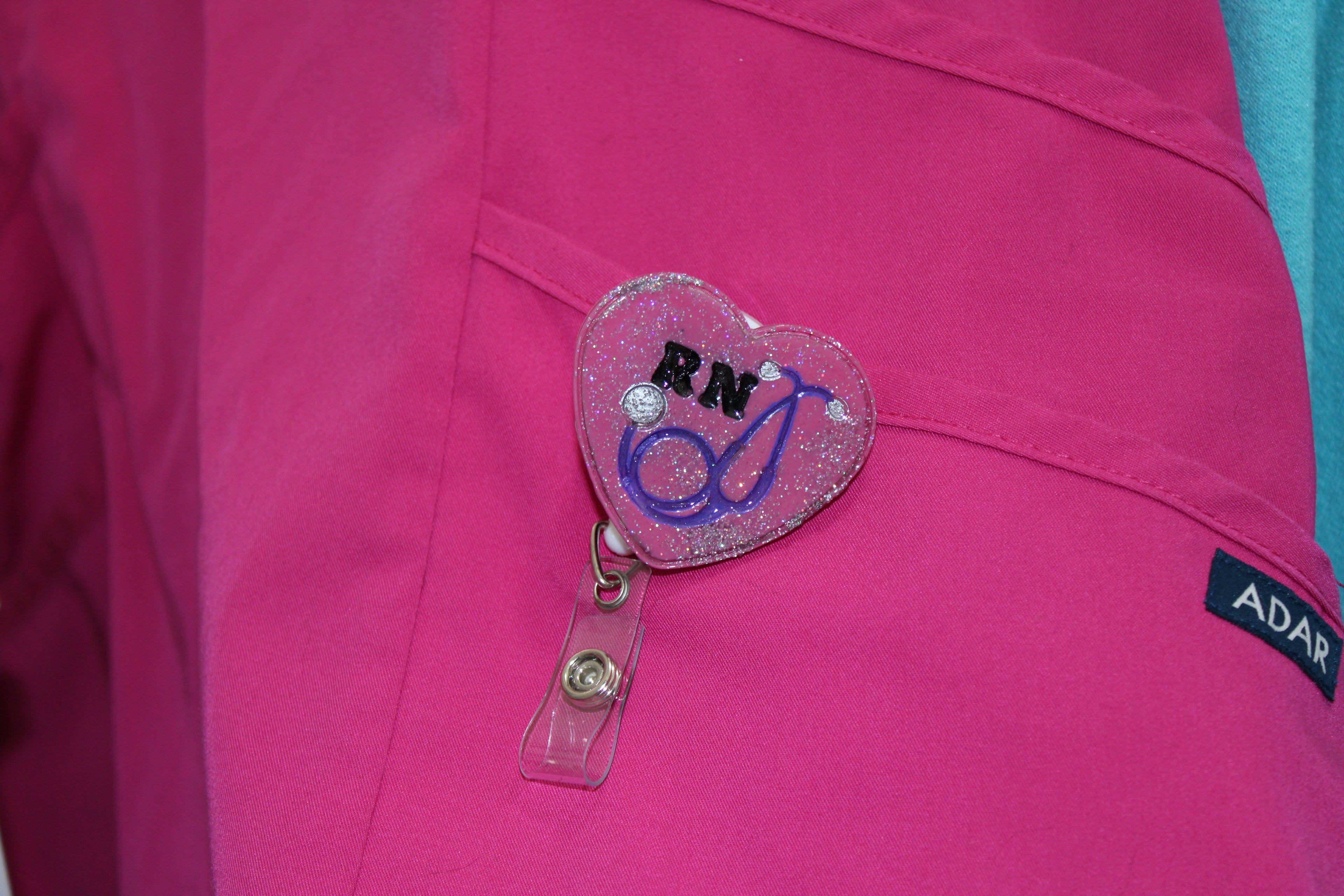 A registered nurse's pink scrub top features a glittery heart-shaped RN badge reel with a stethoscope design.