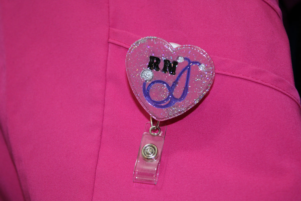 A registered nurse's (RN) pink scrubs display a glittery heart-shaped badge reel featuring "RN" and a stethoscope.
