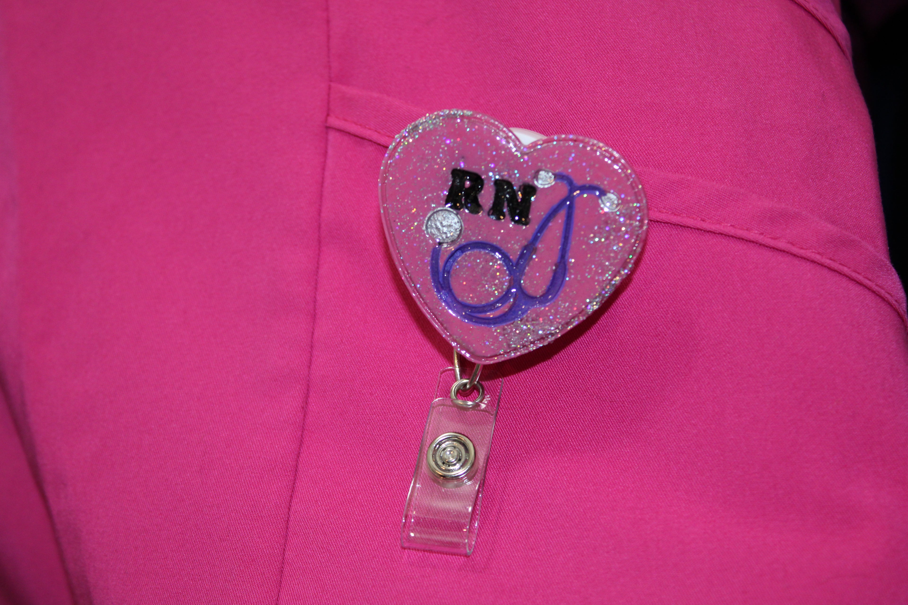 A registered nurse's (RN) pink scrubs display a glittery heart-shaped badge reel featuring "RN" and a stethoscope.