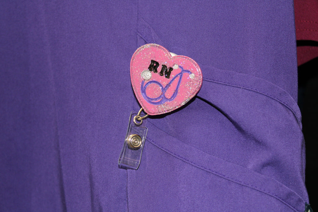 A registered nurse wears a purple scrub top with a sparkly heart-shaped RN badge featuring a stethoscope design.