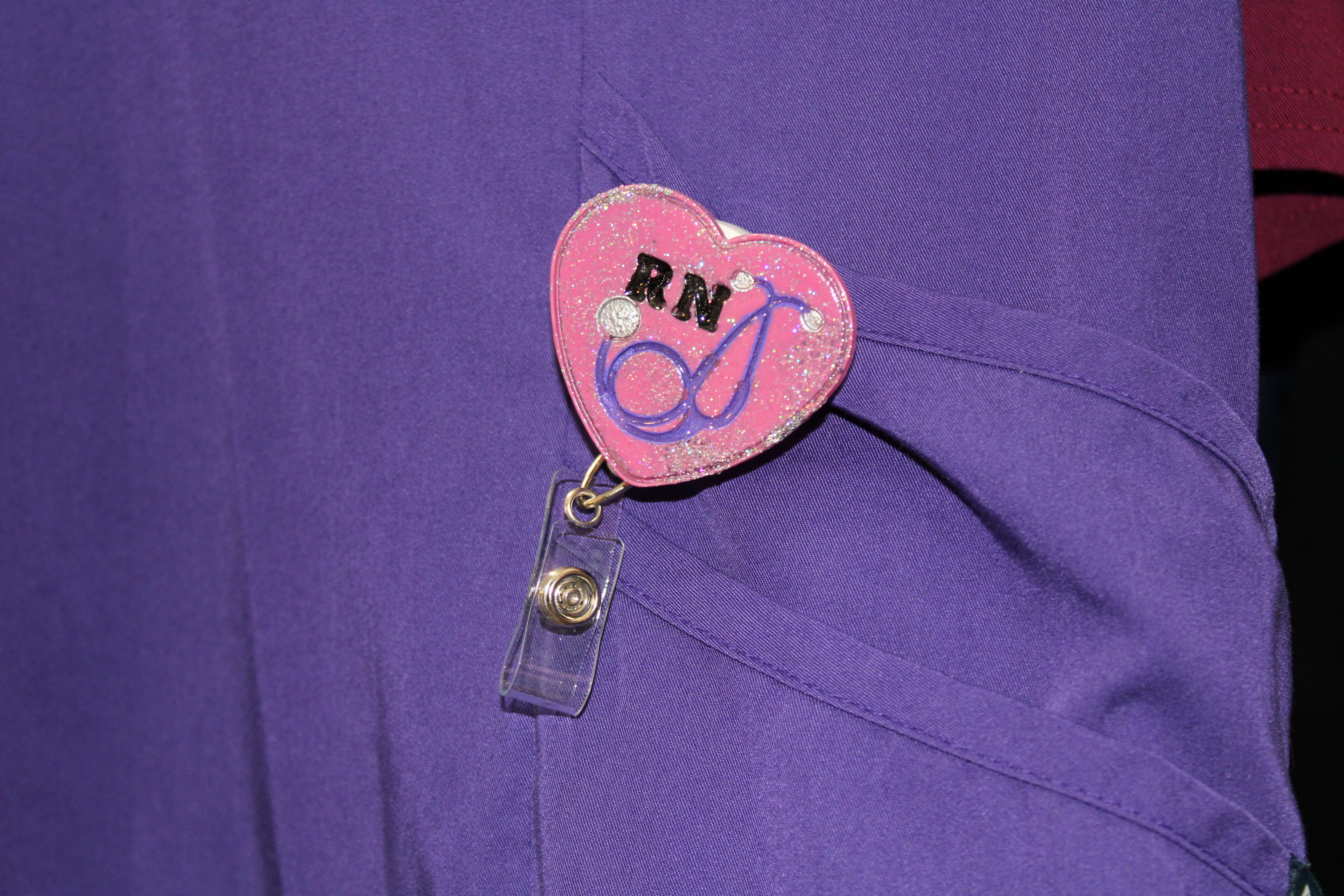 A registered nurse wears a purple scrub top with a sparkly heart-shaped RN badge featuring a stethoscope design.