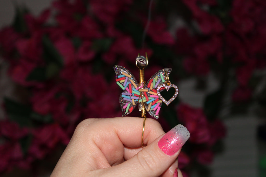 Colorful butterfly-shaped smoke ring held by a hand with pink nail polish against a blurred floral background.