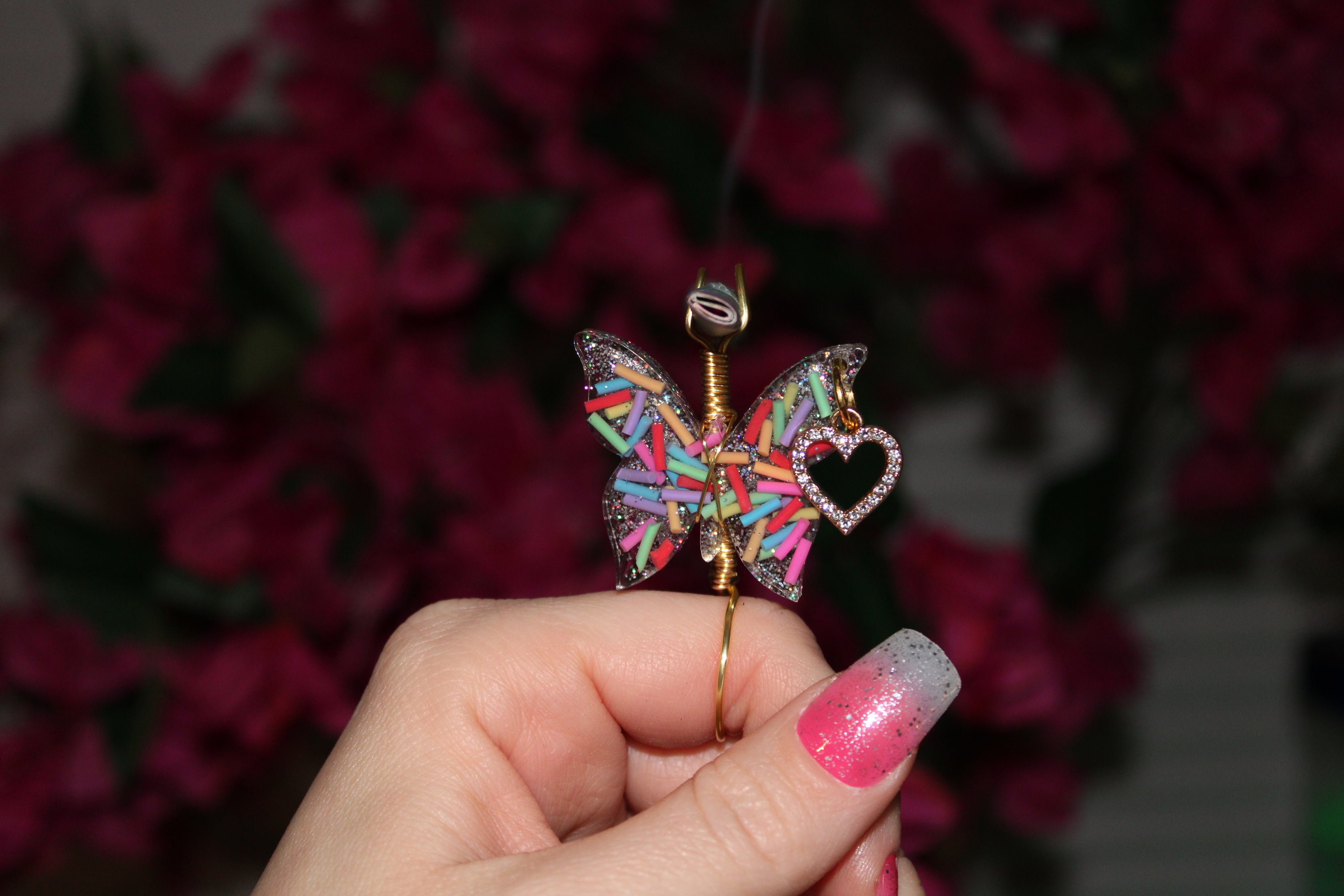 Colorful butterfly-shaped smoke ring held by a hand with pink nail polish against a blurred floral background.