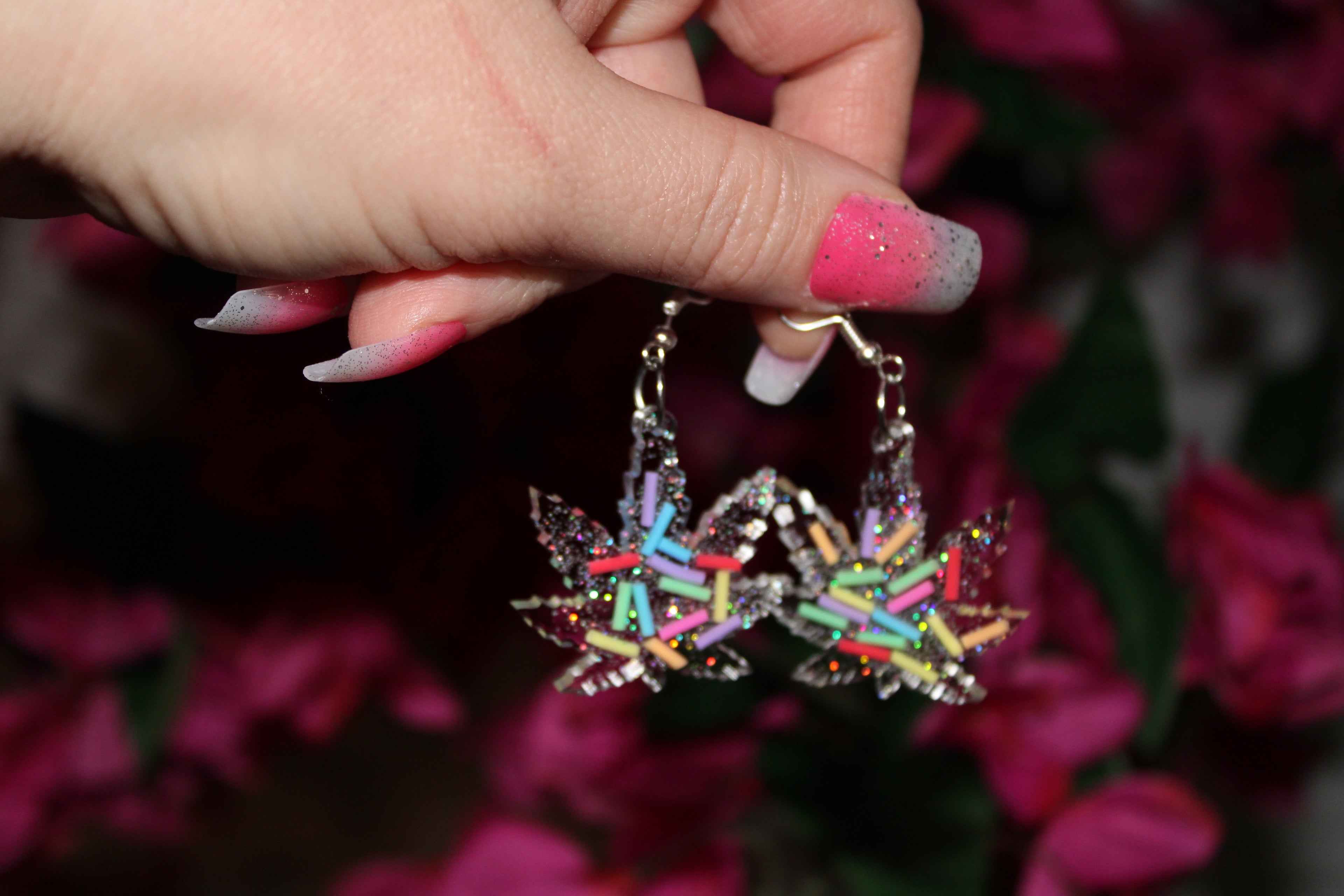 Multicolored marijuana-shaped earrings held by a hand with pink nail polish against a blurred floral background.