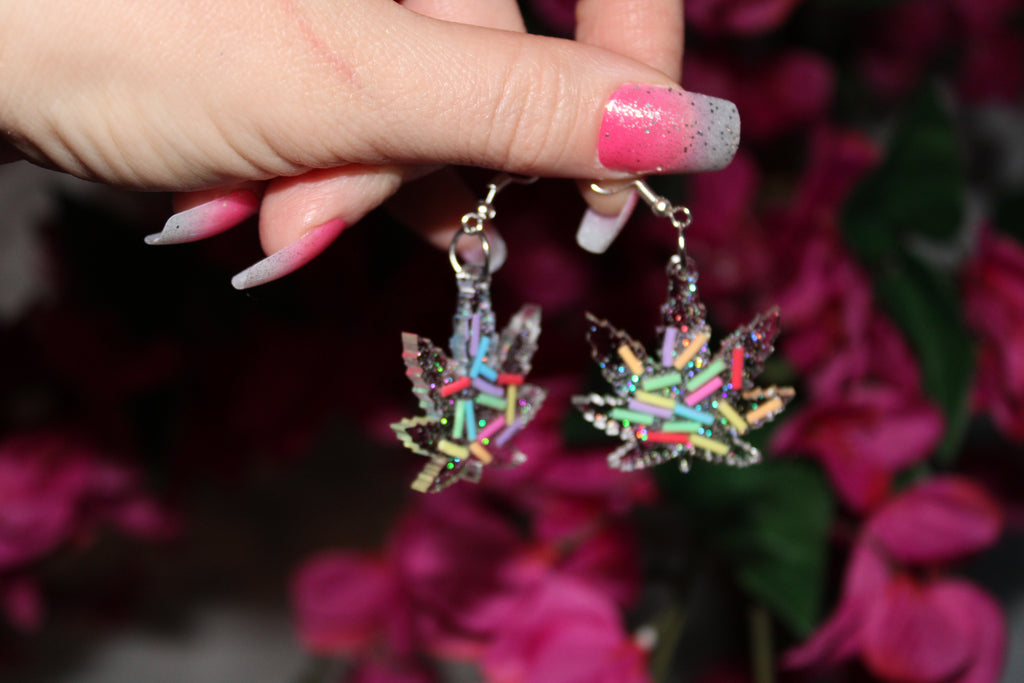 Colorful marijuana leaf-shaped earrings held by a hand with pink nail polish against a blurred floral background.