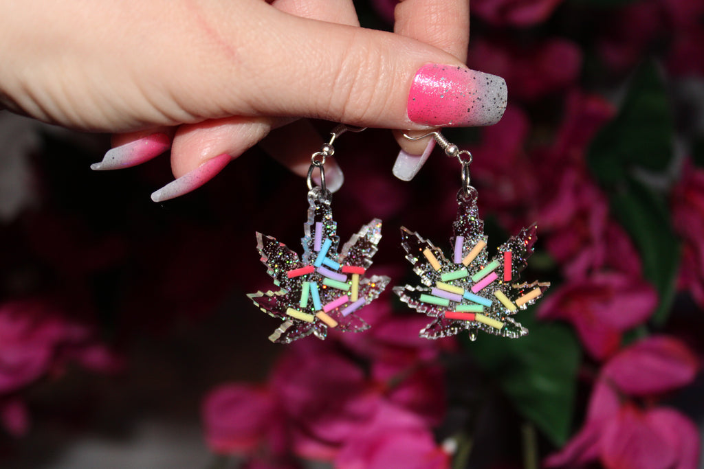 Hand holding marijuana leaf shaped earrings with sprinkle and glitter art inside.