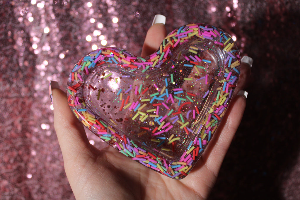 Heart-shaped ashtray with colorful sprinkles held against a glittery pink background