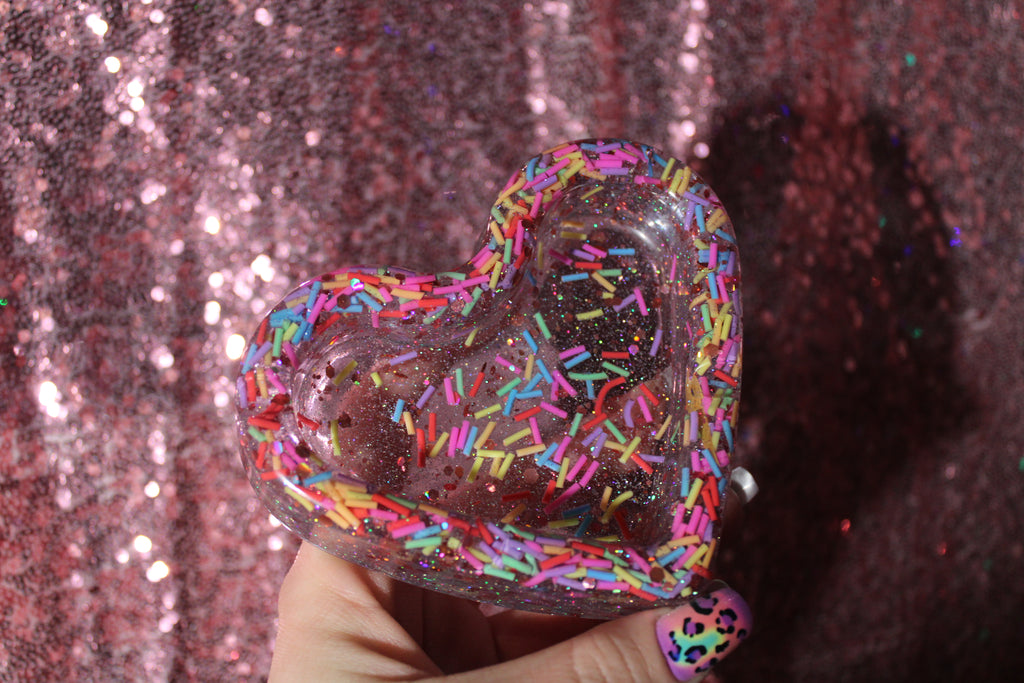 Heart-shaped ashtray with colorful sprinkles against a glittery pink background
