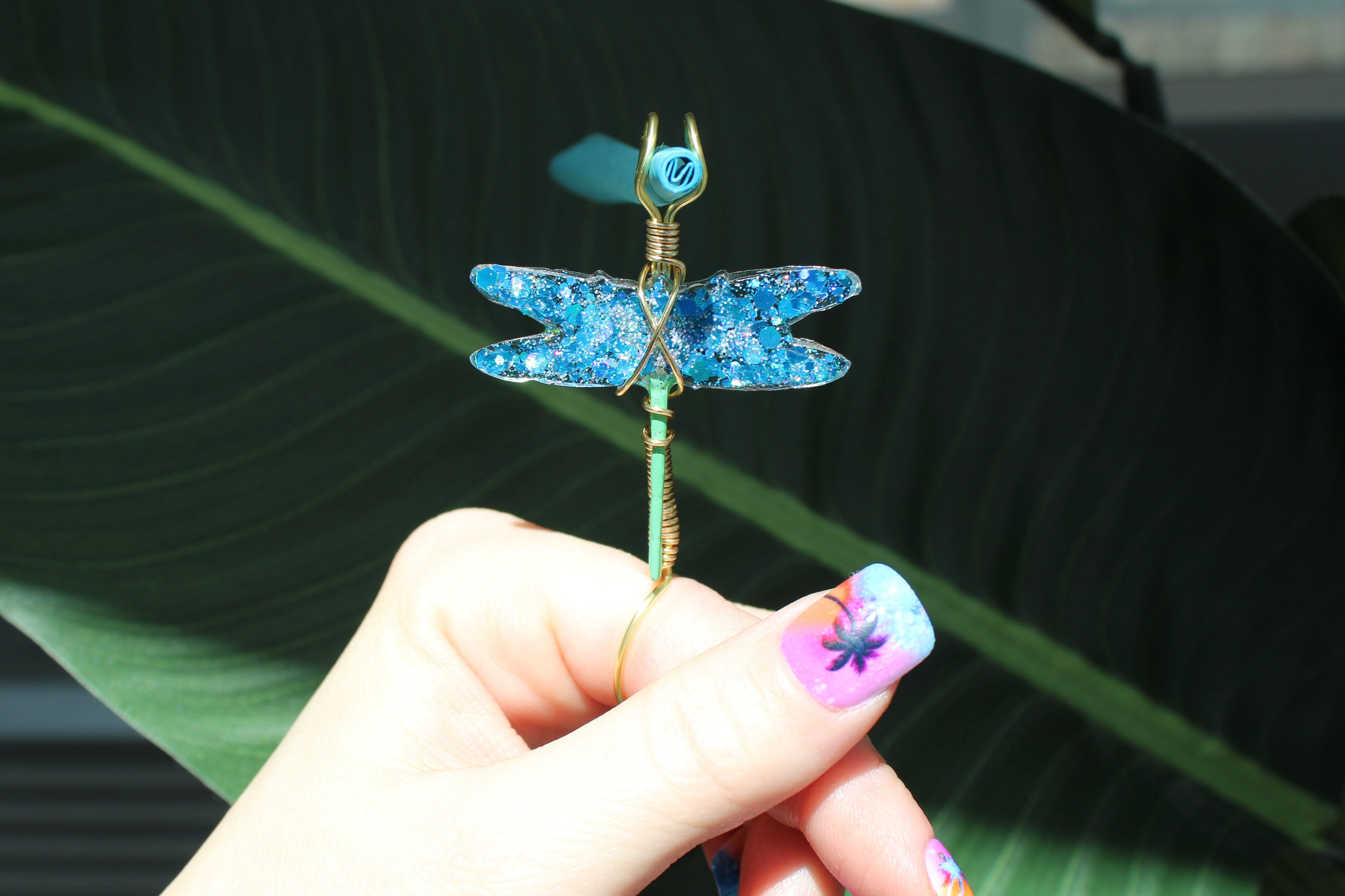Hand holding a decorative dragonfly smoke ring with blue colors against a leafy background.