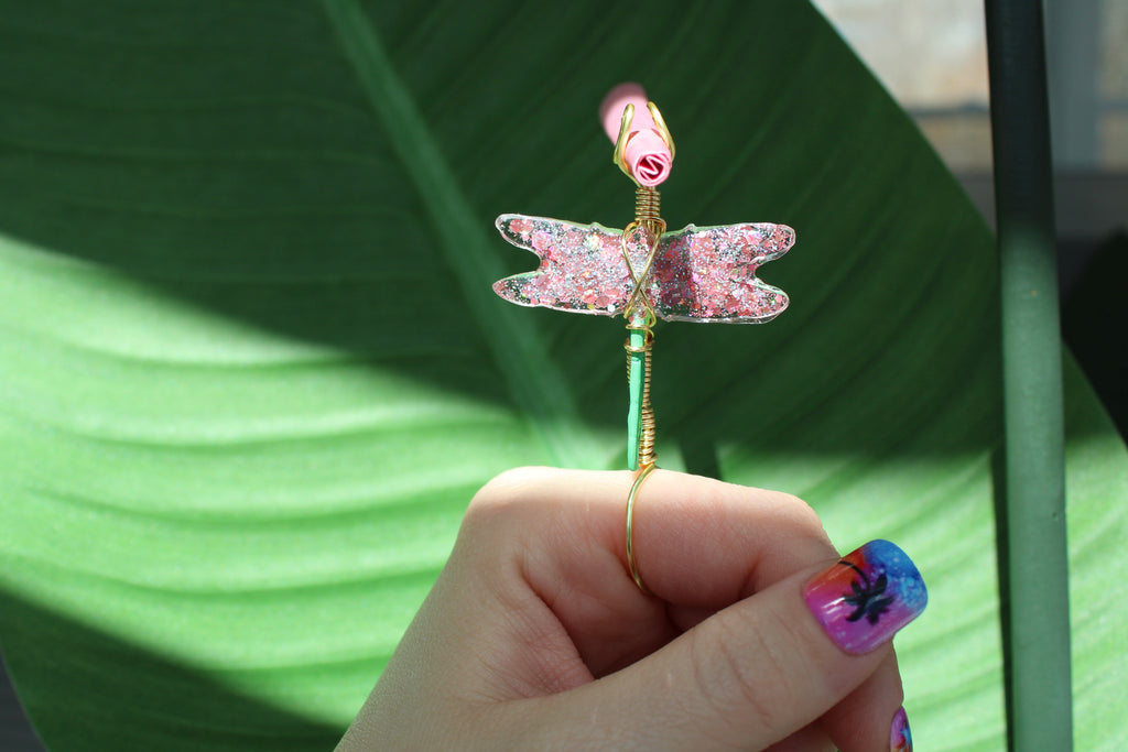 Hand holding a decorative smoke ring shaped like a dragonfly against a green leaf background.