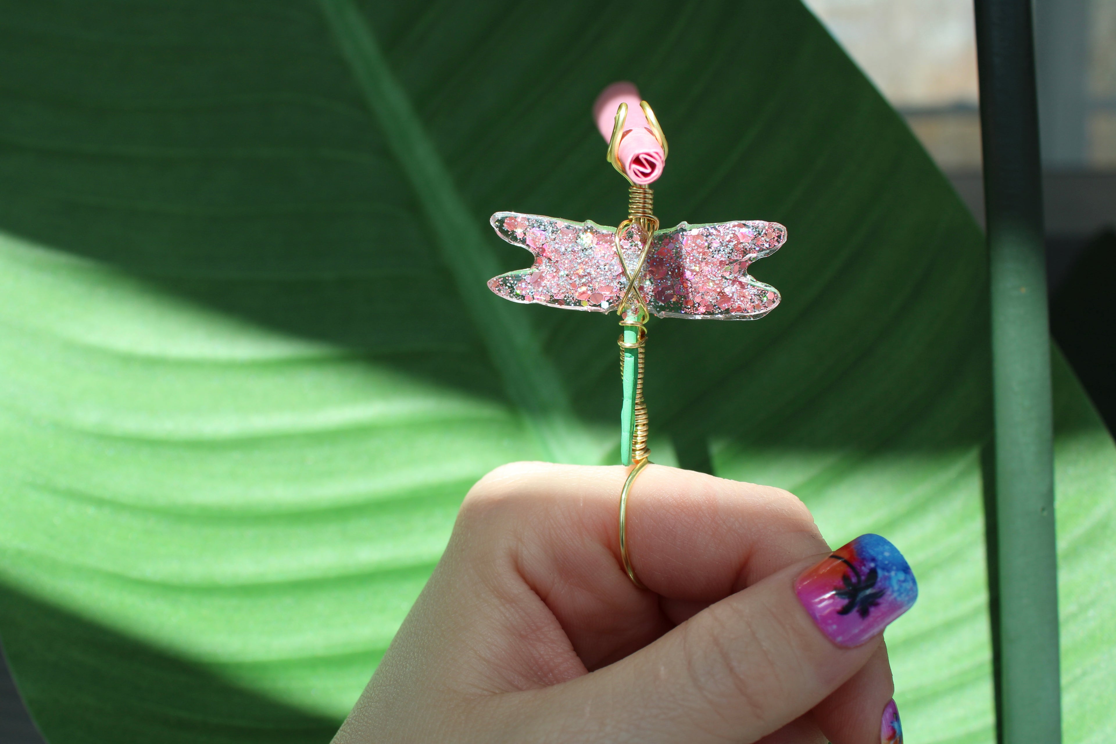 Hand holding a decorative smoke ring shaped like a dragonfly against a green leaf background.