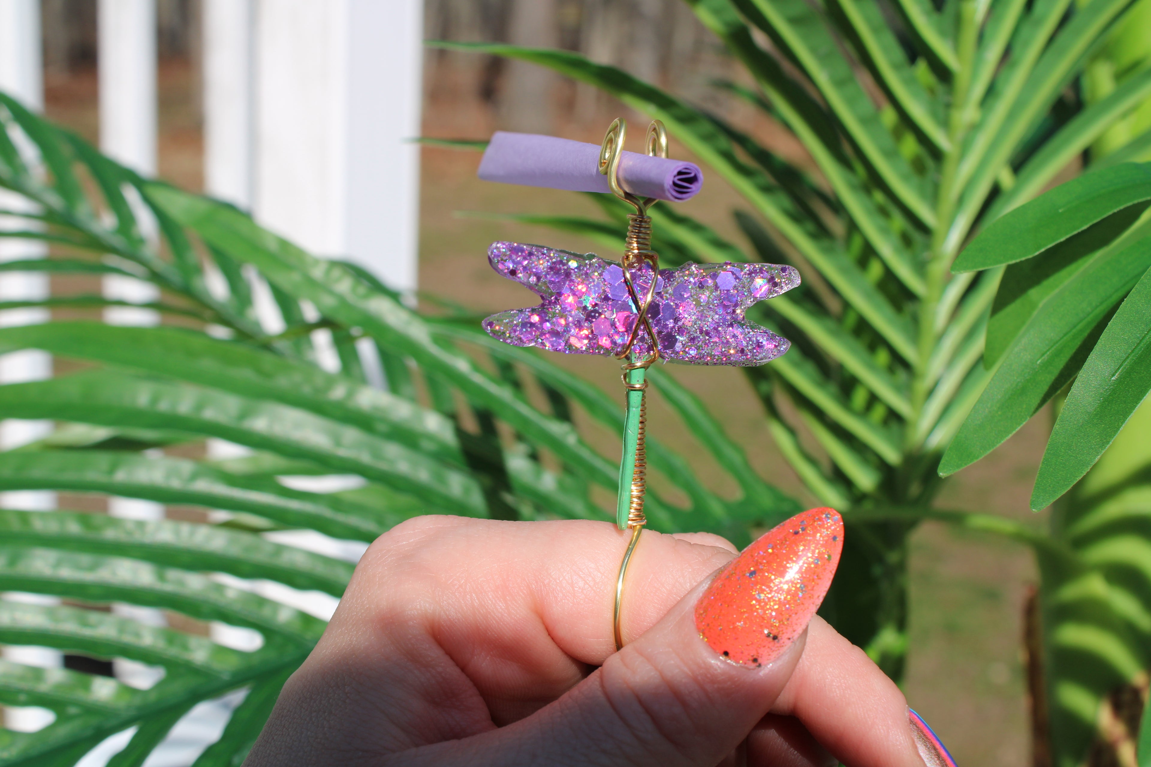 Image shows a purple sequin dragonfly smoke ring with a purple rolling paper in front of a fake plant 