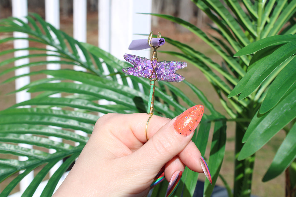 Image shows a purple sequin dragonfly smoke ring with a purple rolling paper in front of a fake plant 