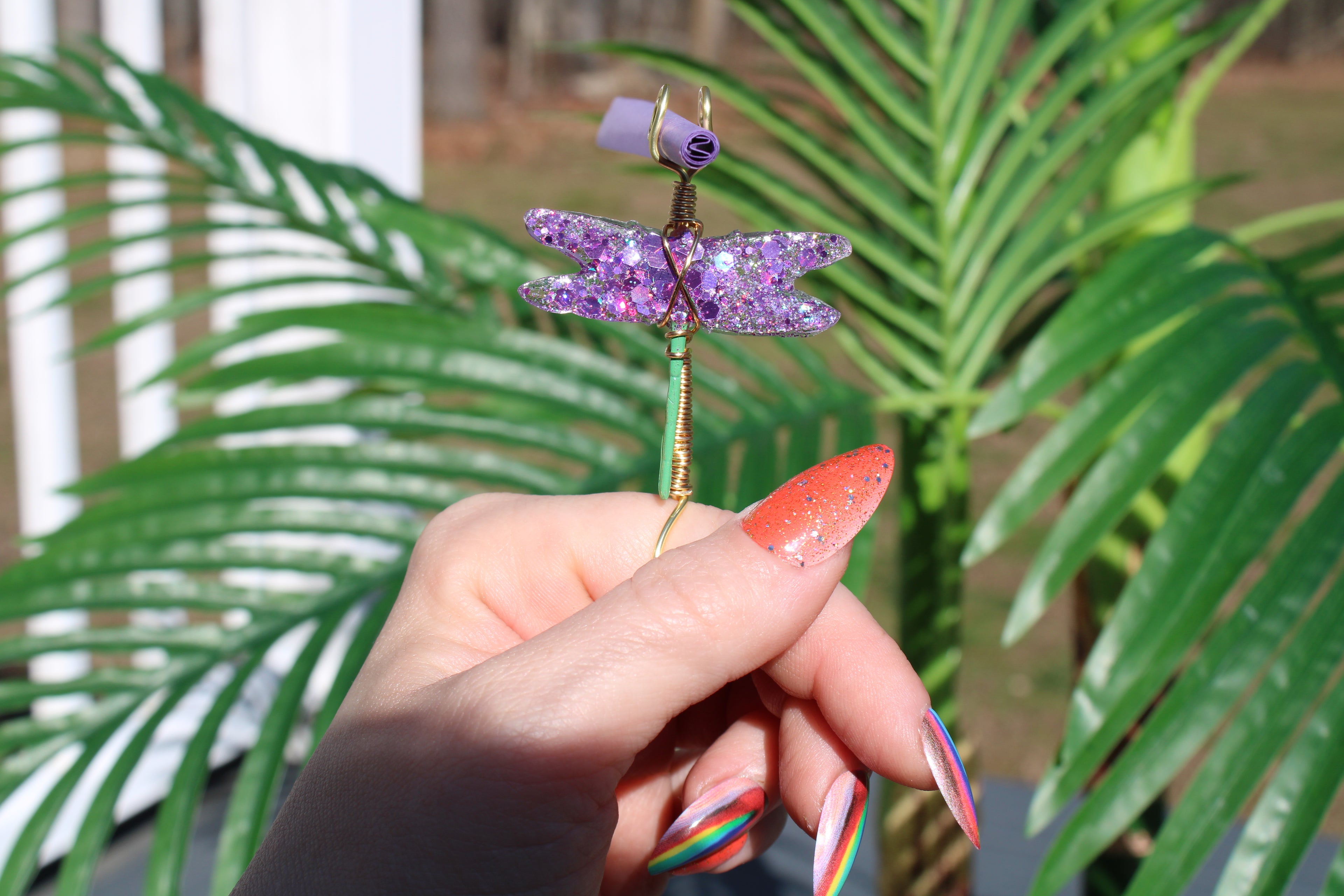 Image shows a purple sequin dragonfly smoke ring with a purple rolling paper in front of a fake plant 